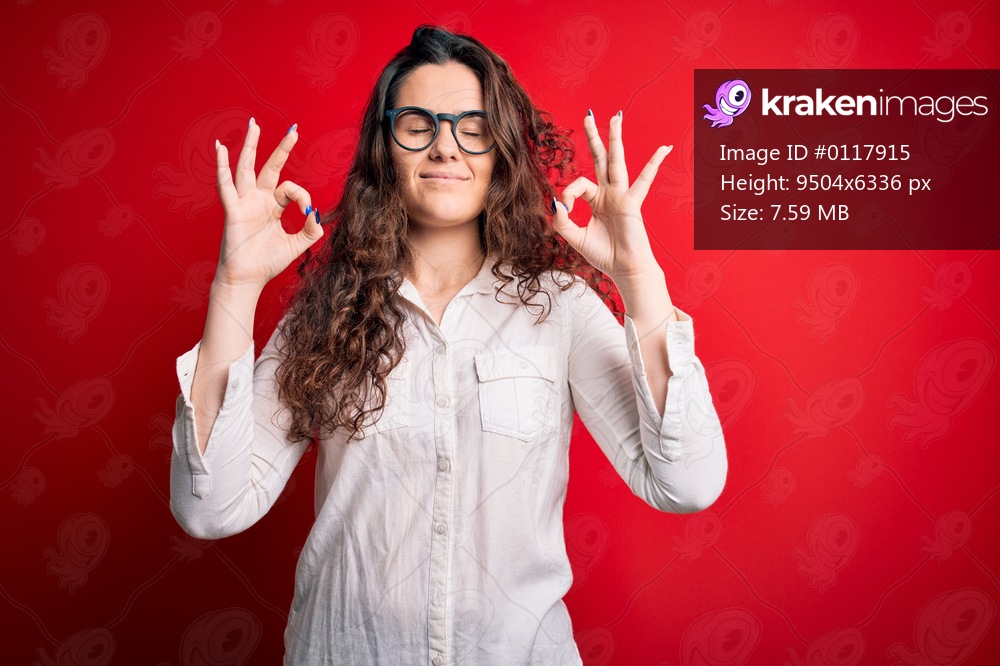 Young beautiful woman with curly hair wearing shirt and glasses over red background relax and smiling with eyes closed doing meditation gesture with fingers. Yoga concept.