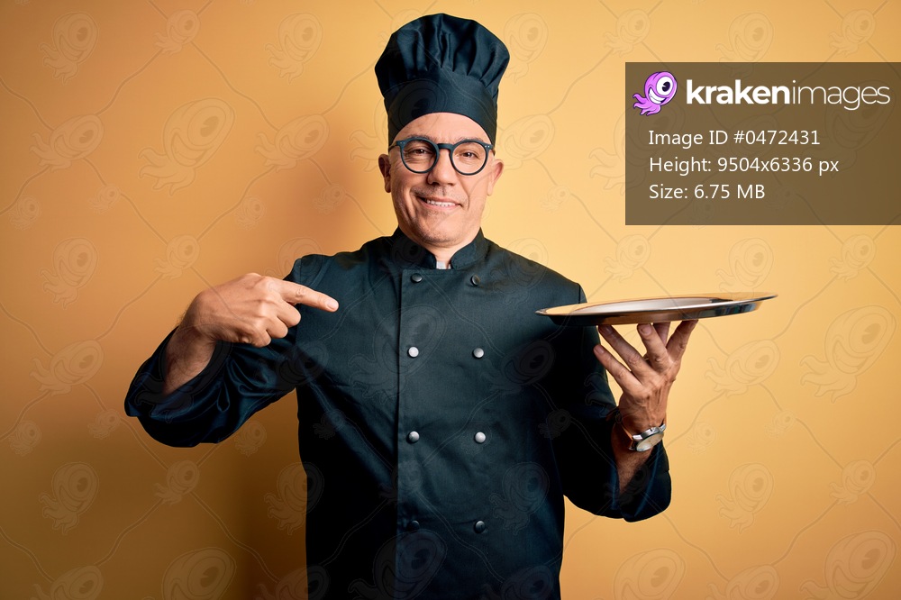 Middle age handsome grey-haired waiter man wearing cooker uniform and hat holding tray with surprise face pointing finger to himself