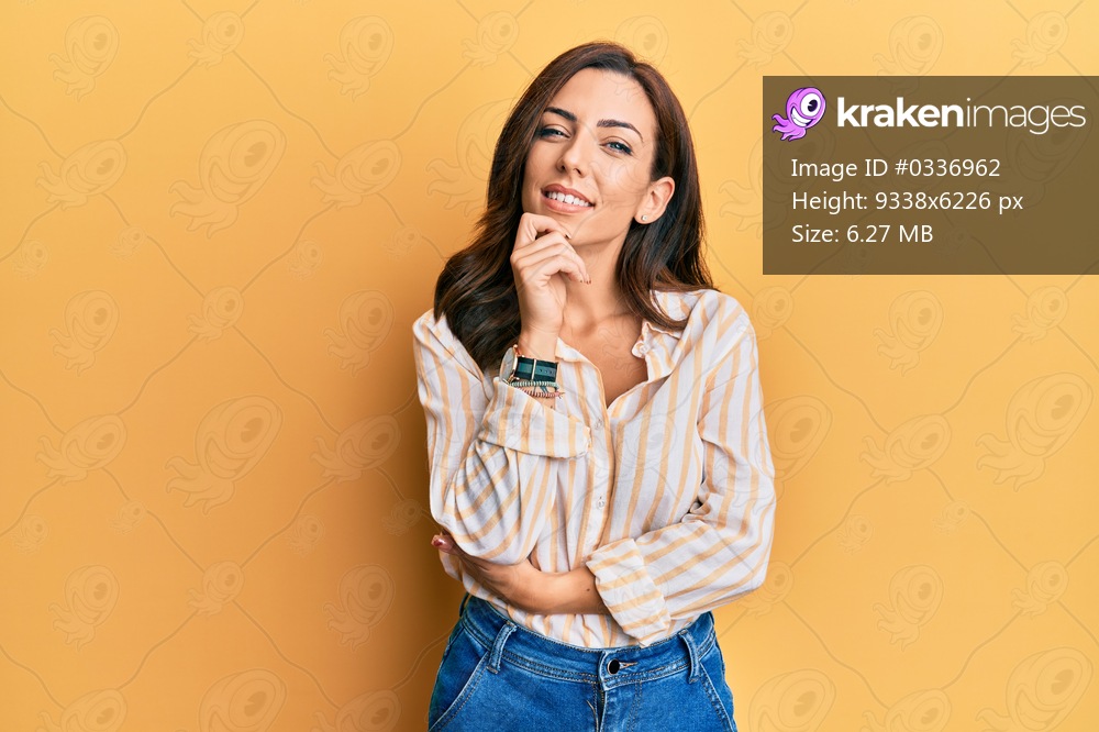 Young brunette woman wearing casual clothes over yellow background smiling looking confident at the camera with crossed arms and hand on chin. thinking positive. 