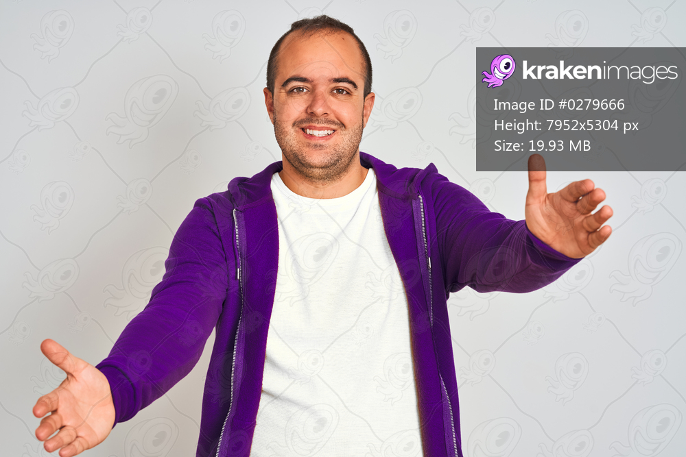Young man wearing purple sweatshirt standing over isolated white background looking at the camera smiling with open arms for hug. Cheerful expression embracing happiness.