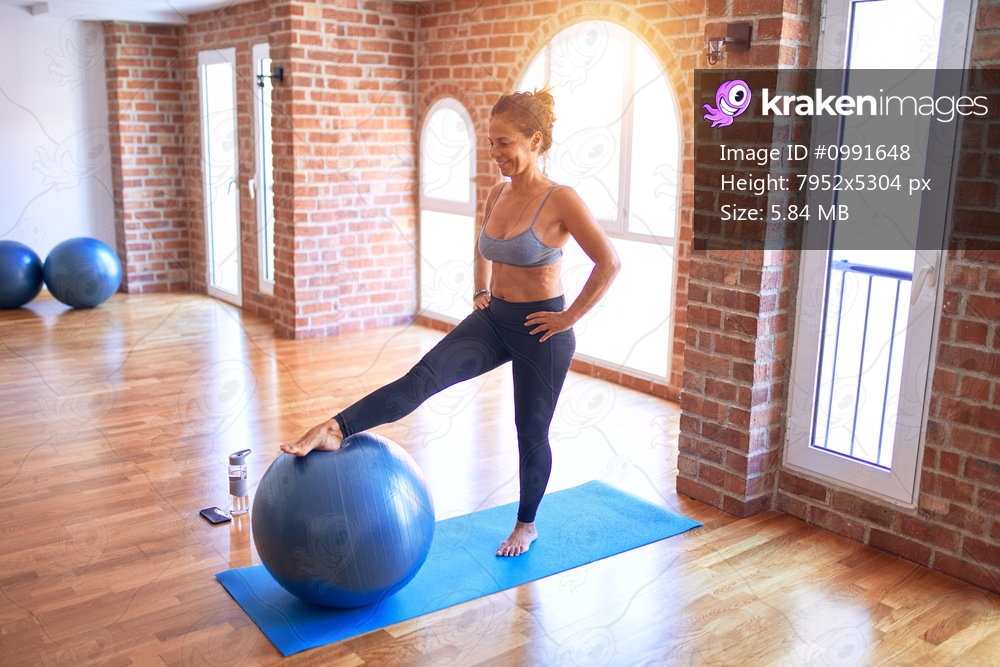 Middle age beautiful sportswoman smiling happy. Doing exercise using fitness ball in a class of yoga at gym