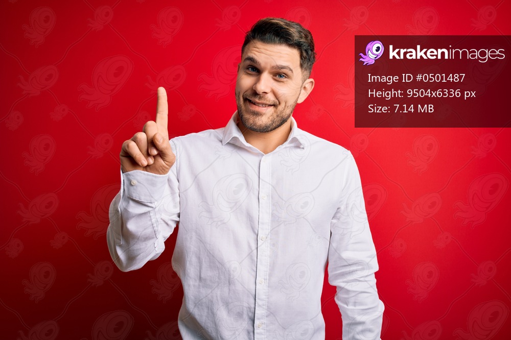 Young business man with blue eyes wearing elegant shirt standing over red isolated background showing and pointing up with finger number one while smiling confident and happy.