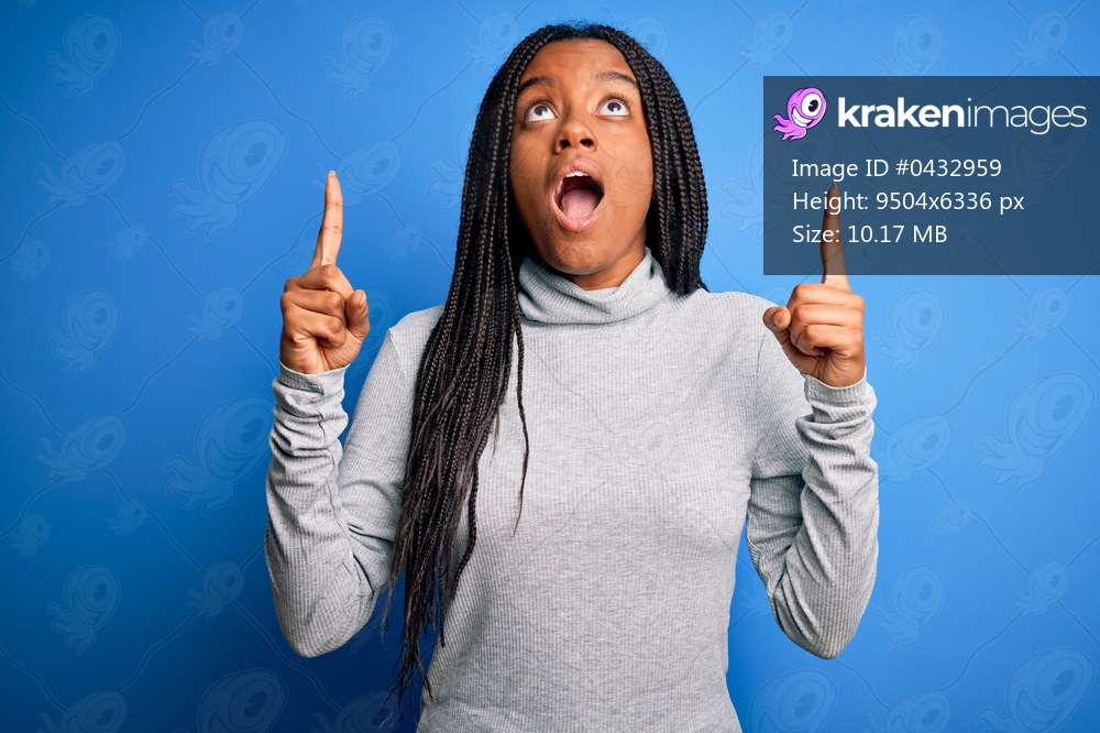 Young african american woman standing wearing casual turtleneck over blue isolated background amazed and surprised looking up and pointing with fingers and raised arms.