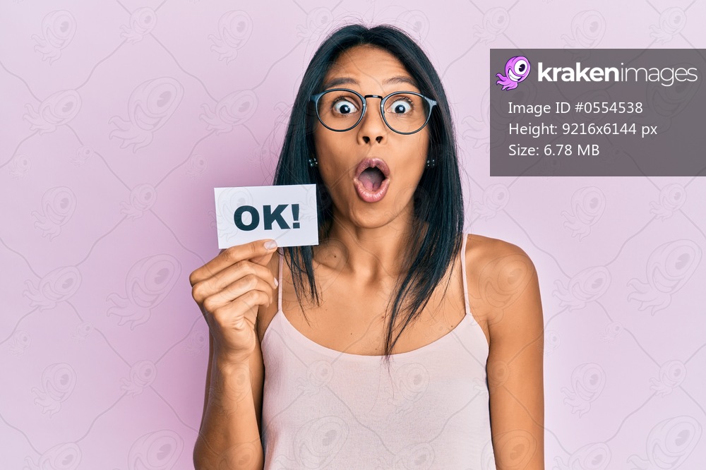 Young african american woman holding ok message paper scared and amazed with open mouth for surprise, disbelief face 