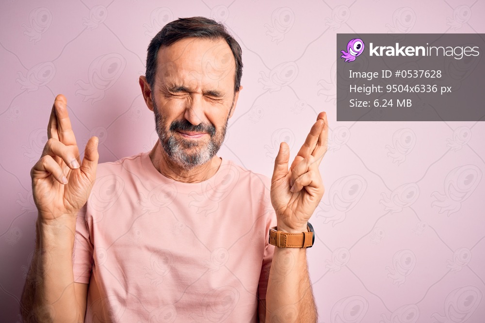 Middle age hoary man wearing casual t-shirt standing over isolated pink background gesturing finger crossed smiling with hope and eyes closed. Luck and superstitious concept.