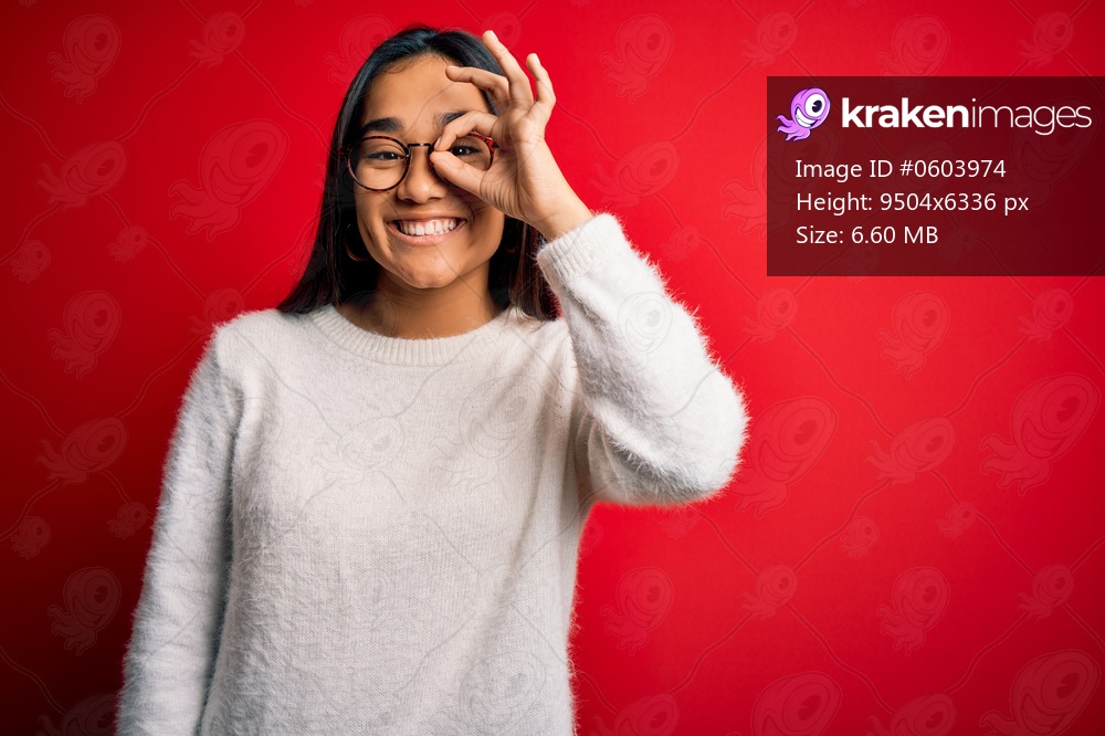 Young beautiful asian woman wearing casual sweater and glasses over red background doing ok gesture with hand smiling, eye looking through fingers with happy face.