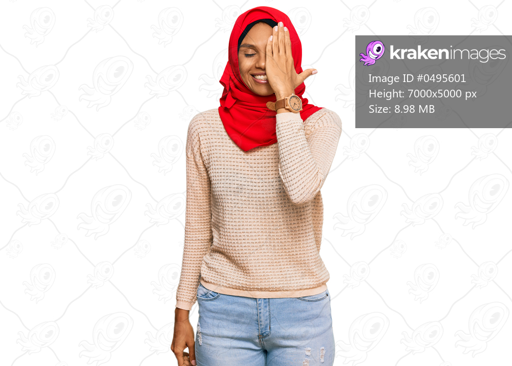 Young african american woman wearing traditional islamic hijab scarf covering one eye with hand, confident smile on face and surprise emotion. 