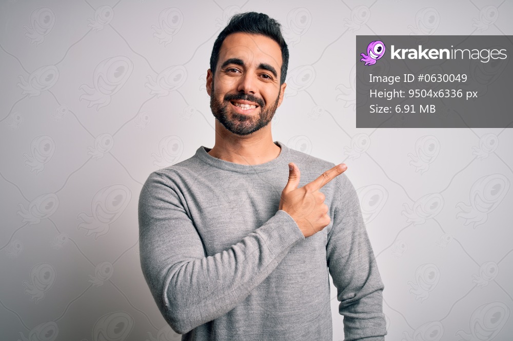 Young handsome man with beard wearing casual sweater standing over white background cheerful with a smile of face pointing with hand and finger up to the side with happy and natural expression on face