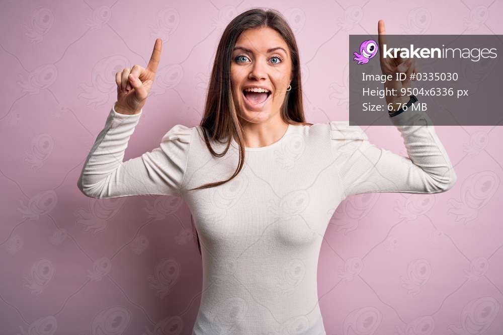 Young beautiful woman with blue eyes wearing casual white t-shirt over pink background smiling amazed and surprised and pointing up with fingers and raised arms.
