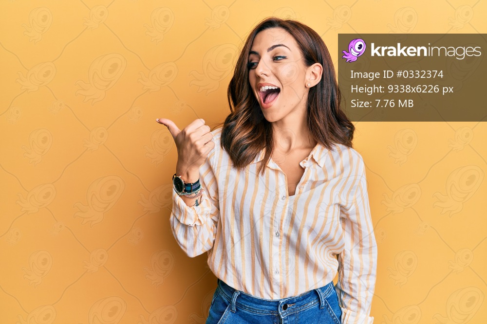 Young brunette woman wearing casual clothes over yellow background pointing thumb up to the side smiling happy with open mouth 