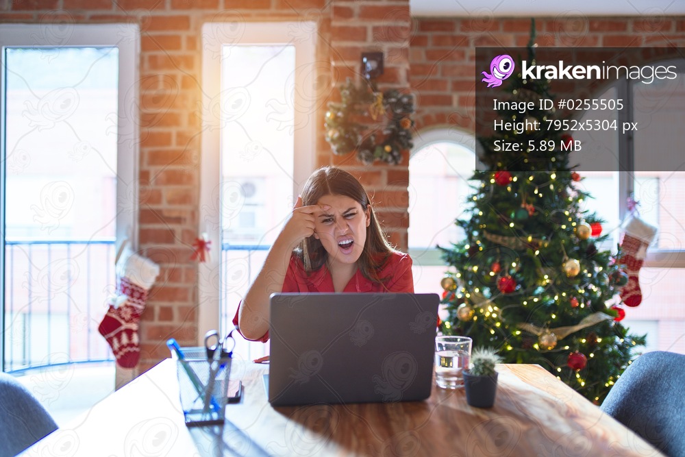 Beautiful woman sitting at the table working with laptop at home around christmas tree pointing unhappy to pimple on forehead, ugly infection of blackhead. Acne and skin problem