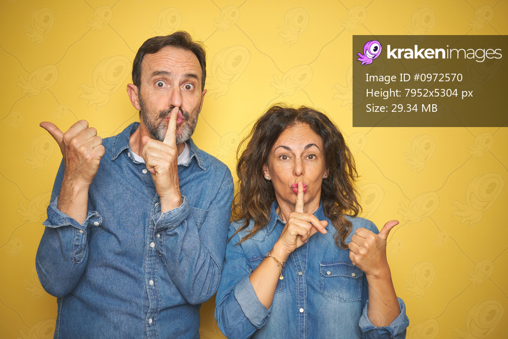 Beautiful middle age couple together standing over isolated yellow background asking to be quiet with finger on lips pointing with hand to the side. Silence and secret concept.