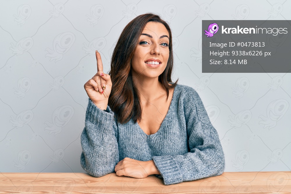 Young brunette woman wearing casual sweater sitting on the table smiling with an idea or question pointing finger up with happy face, number one 