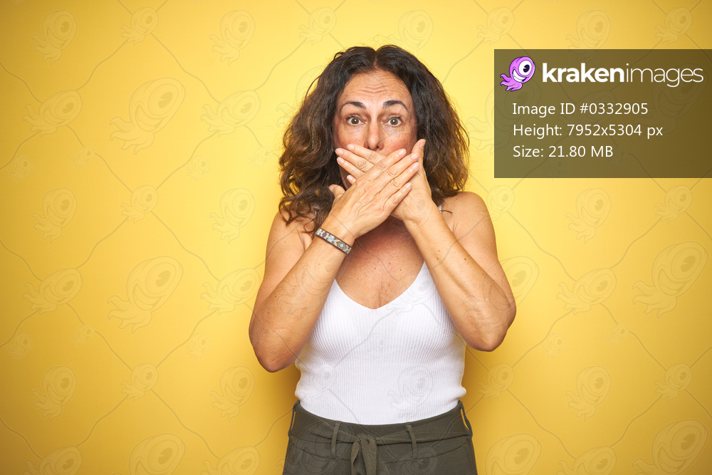 Middle age senior woman with curly hair standing over yellow isolated background shocked covering mouth with hands for mistake. Secret concept.