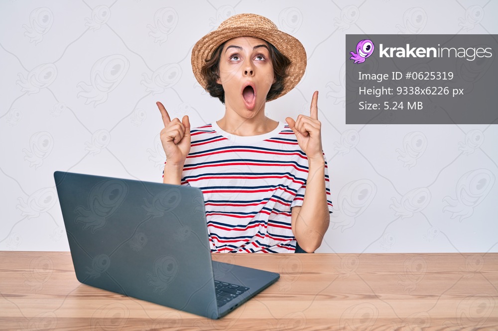 Young hispanic woman working with laptop sitting on the table amazed and surprised looking up and pointing with fingers and raised arms. 