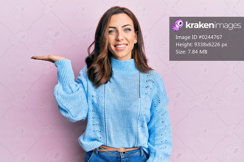 Young brunette woman wearing casual winter sweater over pink background smiling cheerful presenting and pointing with palm of hand looking at the camera. 