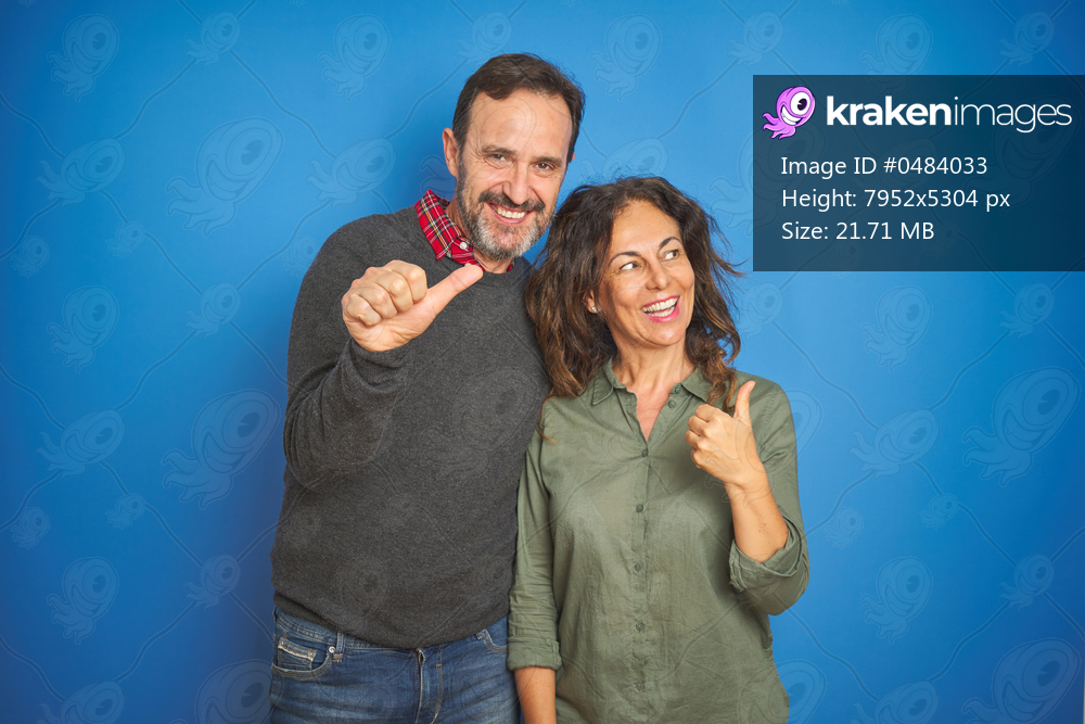 Beautiful middle age couple together standing over isolated blue background smiling with happy face looking and pointing to the side with thumb up.