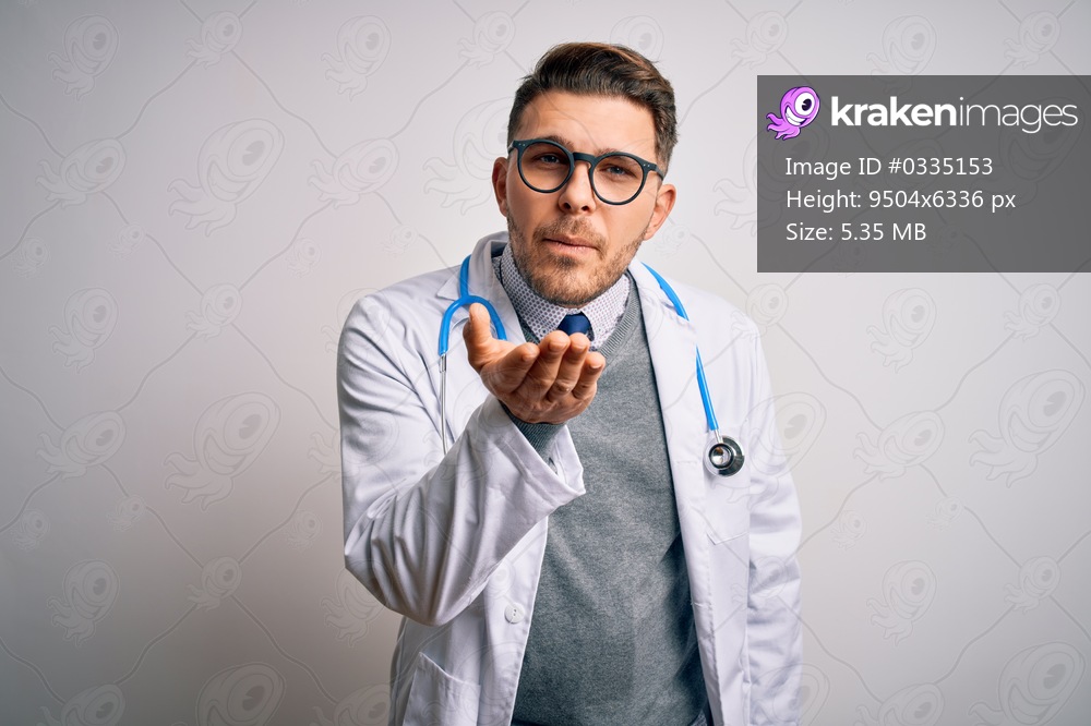 Young doctor man with blue eyes wearing medical coat and stethoscope over isolated background looking at the camera blowing a kiss with hand on air being lovely and sexy. Love expression.