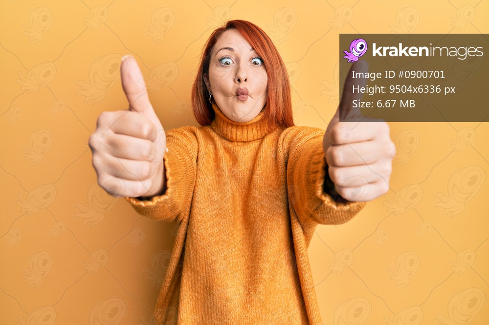 Beautiful redhead woman doing thumbs up positive gesture making fish face with mouth and squinting eyes, crazy and comical. 