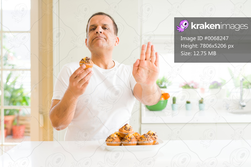 Middle age man eating chocolate chip muffins at home with open hand doing stop sign with serious and confident expression, defense gesture