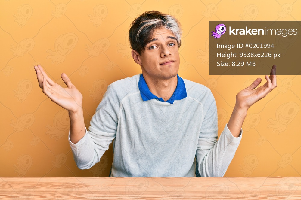 Young hispanic man wearing casual clothes sitting on the table clueless and confused with open arms, no idea and doubtful face. 