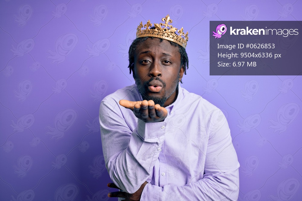 Young african american man wearing golden crown of king over isolated purple background looking at the camera blowing a kiss with hand on air being lovely and sexy. Love expression.