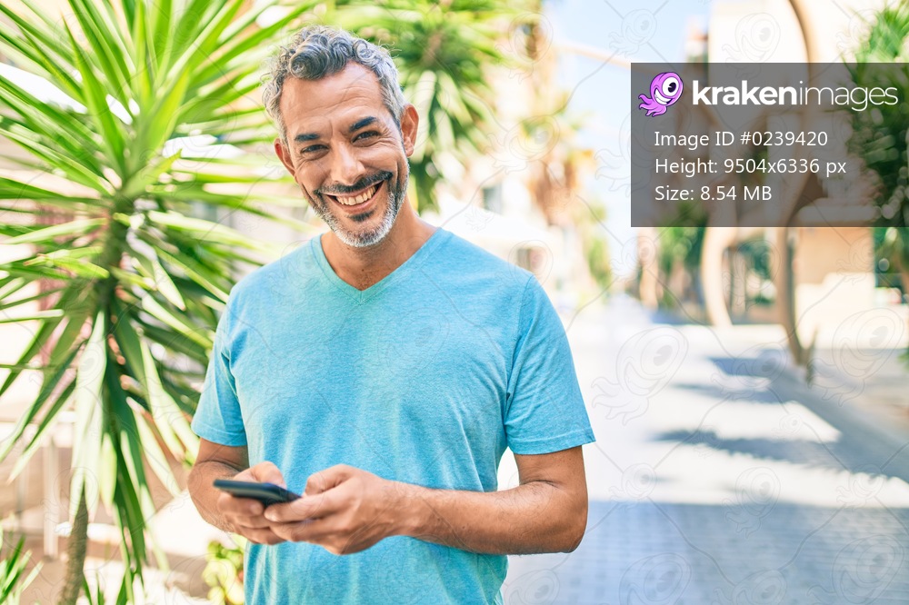 Middle age grey-haired man smiling happy using smartphone walking at street of city.