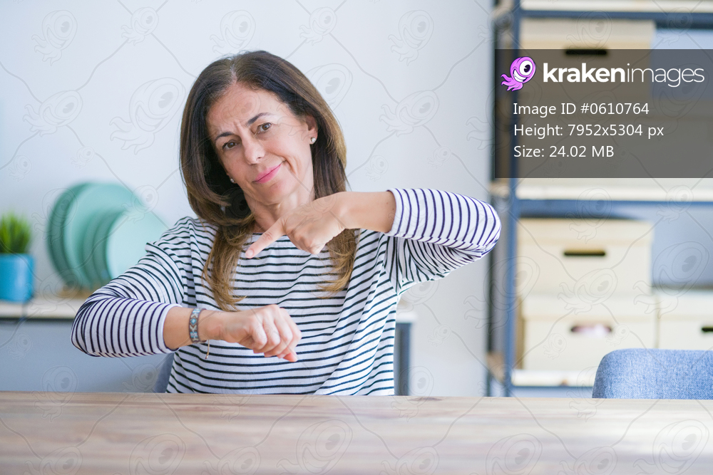 Middle age senior woman sitting at the table at home In hurry pointing to watch time, impatience, upset and angry for deadline delay