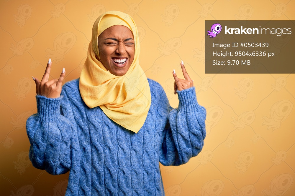 Young African American afro woman wearing muslim hijab over isolated yellow background shouting with crazy expression doing rock symbol with hands up. Music star. Heavy concept.
