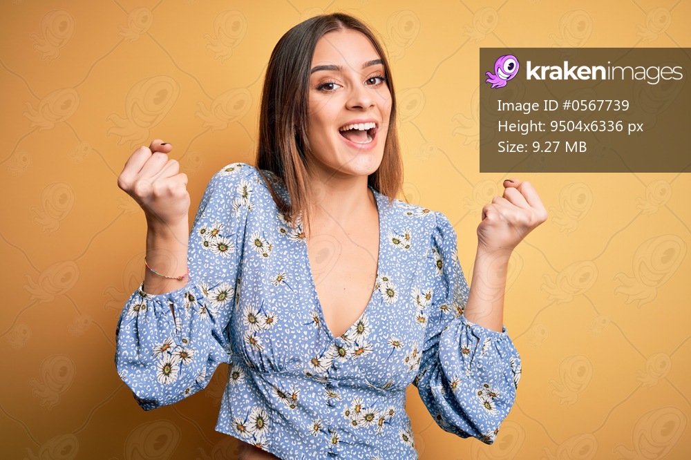 Young beautiful brunette woman wearing casual floral t-shirt standing over yellow background celebrating surprised and amazed for success with arms raised and open eyes. Winner concept.