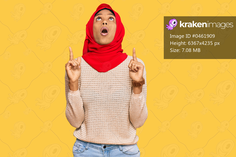 Young african american woman wearing traditional islamic hijab scarf amazed and surprised looking up and pointing with fingers and raised arms. 