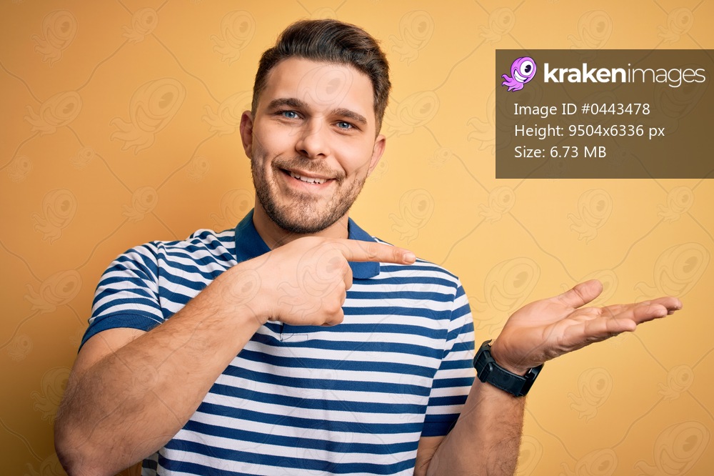 Young man with blue eyes wearing casual striped t-shirt over yellow background amazed and smiling to the camera while presenting with hand and pointing with finger.