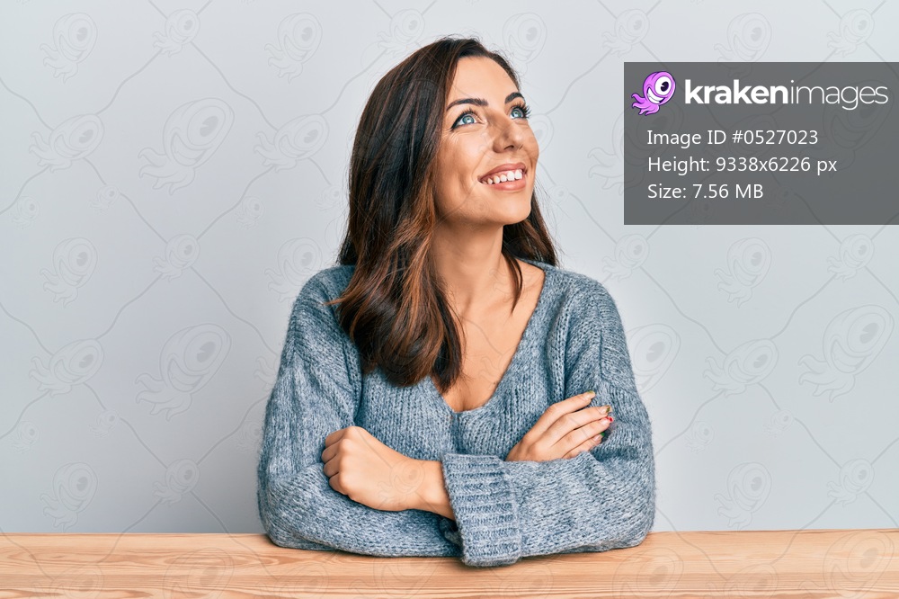 Young brunette woman wearing casual sweater sitting on the table looking to side, relax profile pose with natural face and confident smile. 