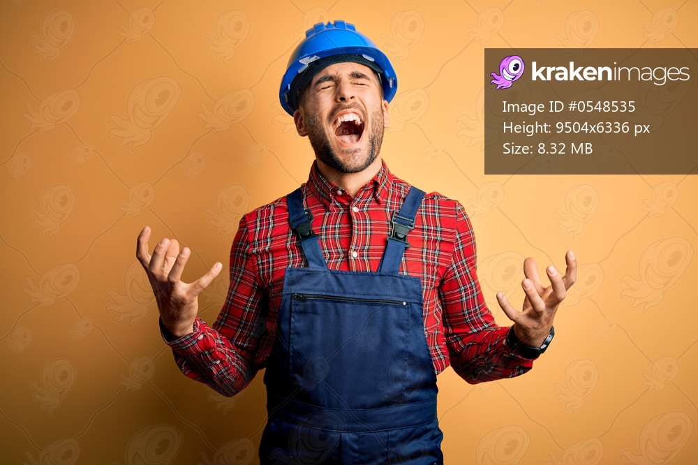 Young builder man wearing construction uniform and safety helmet over yellow background crazy and mad shouting and yelling with aggressive expression and arms raised. Frustration concept.