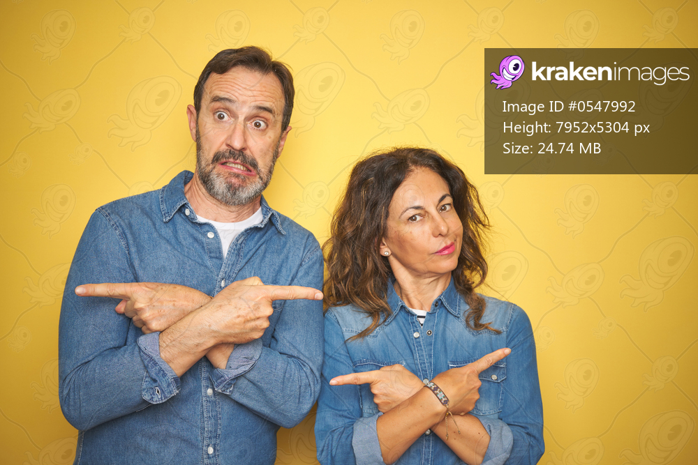 Beautiful middle age couple together standing over isolated yellow background Pointing to both sides with fingers, different direction disagree