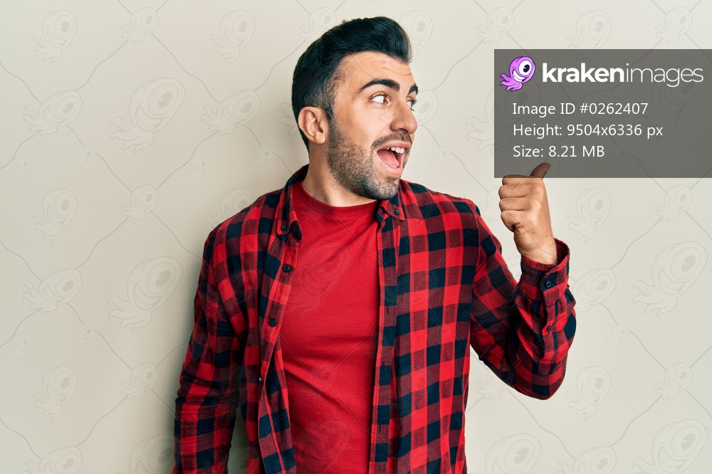 Young hispanic man wearing casual clothes pointing thumb up to the side smiling happy with open mouth 