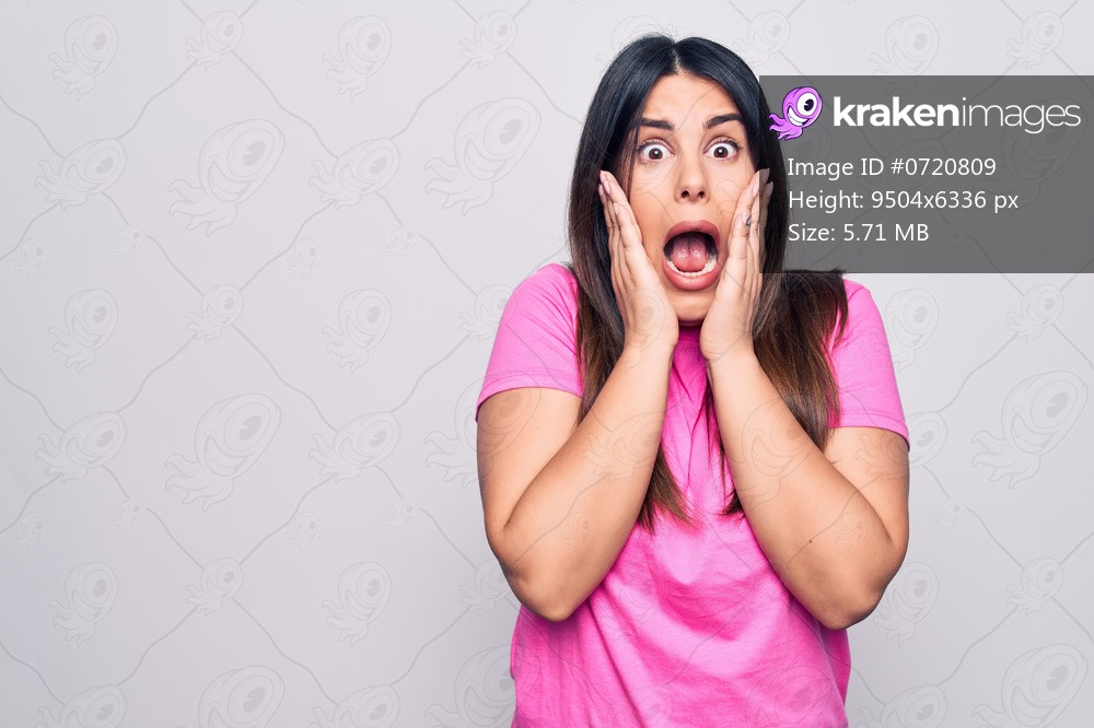 Young beautiful brunette woman wearing casual pink t-shirt standing over white background afraid and shocked, surprise and amazed expression with hands on face