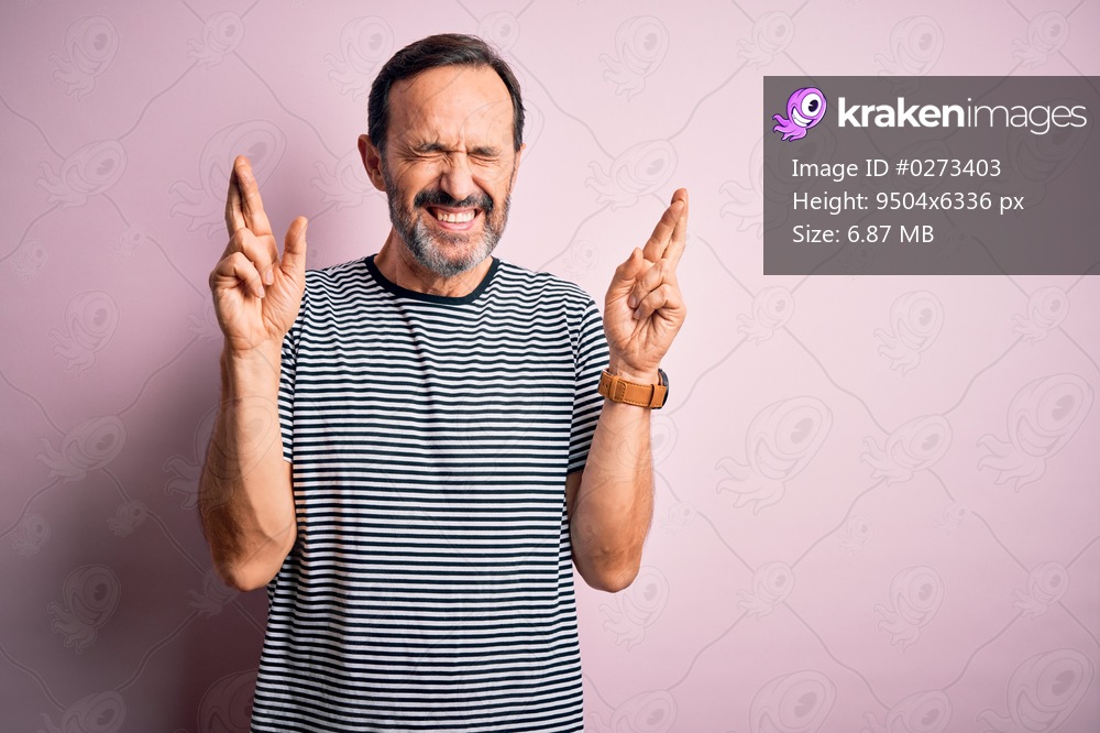 Middle age hoary man wearing casual striped t-shirt standing over isolated pink background gesturing finger crossed smiling with hope and eyes closed. Luck and superstitious concept.