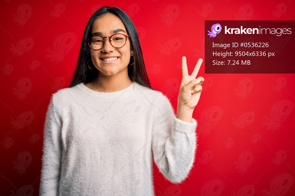 Young beautiful asian woman wearing casual sweater and glasses over red background smiling looking to the camera showing fingers doing victory sign. Number two.