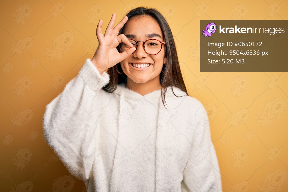 Young beautiful asian woman wearing casual sweater and glasses over yellow background doing ok gesture with hand smiling, eye looking through fingers with happy face.