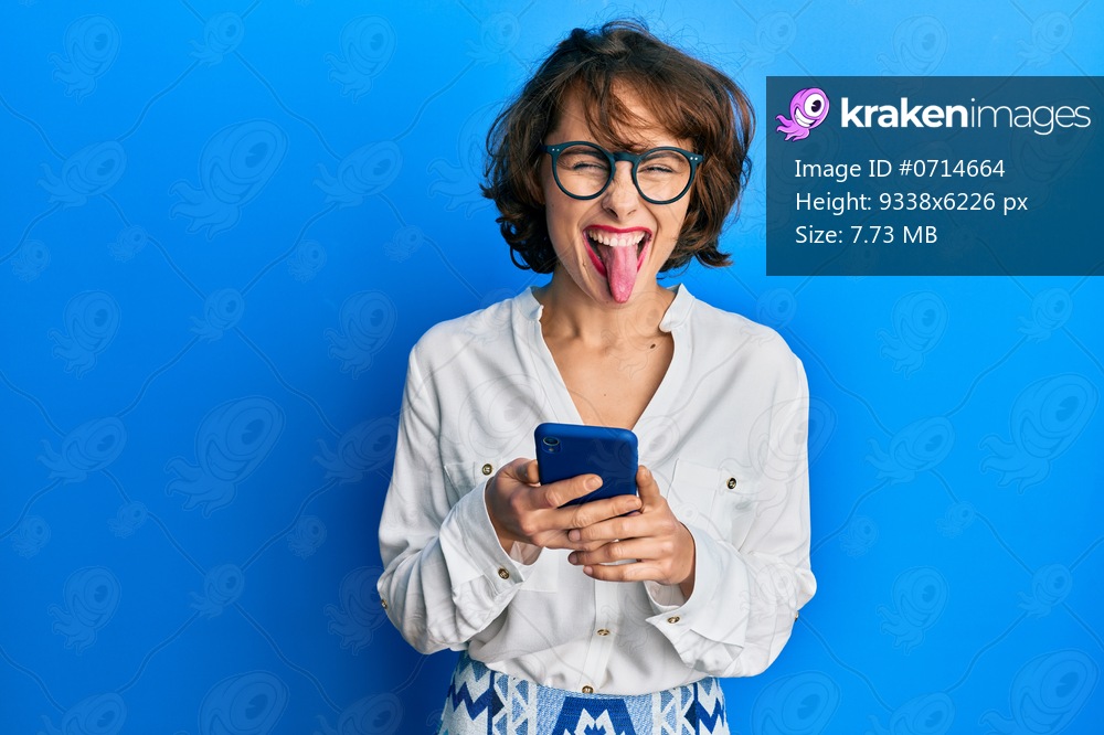 Young brunette woman using smartphone sticking tongue out happy with funny expression. 