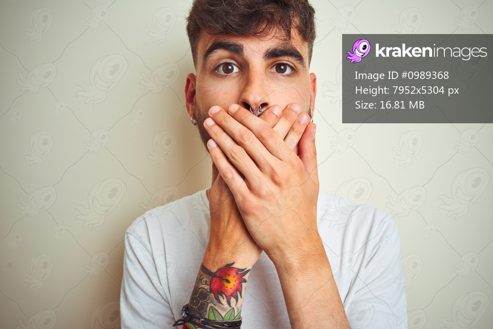 Young man with tattoo wearing t-shirt standing over isolated white background shocked covering mouth with hands for mistake. Secret concept.