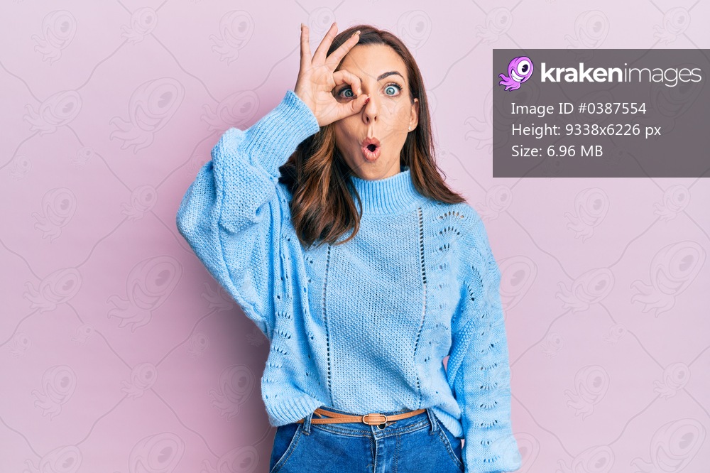 Young brunette woman wearing casual winter sweater over pink background doing ok gesture shocked with surprised face, eye looking through fingers. unbelieving expression. 
