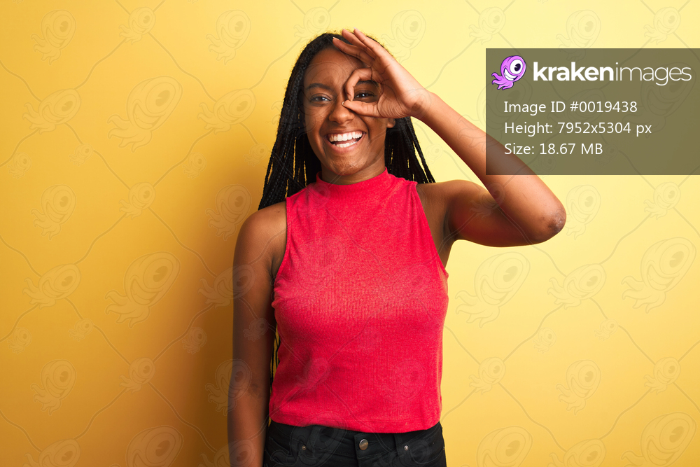 African american woman wearing red casual t-shirt standing over isolated yellow background doing ok gesture with hand smiling, eye looking through fingers with happy face.