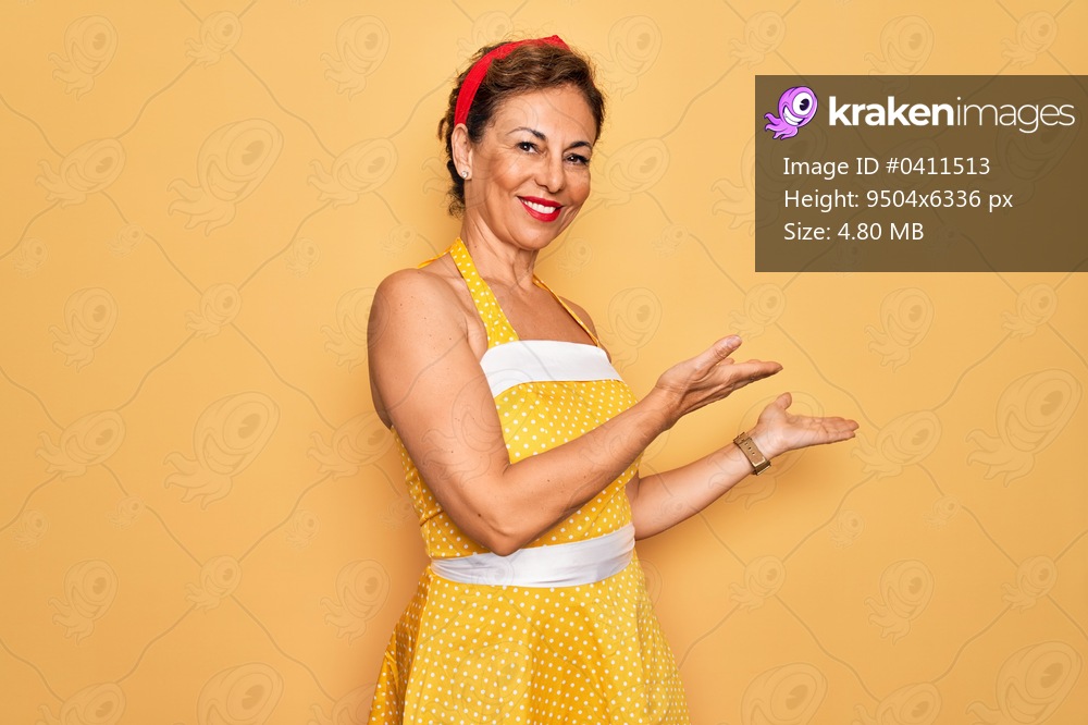 Middle age senior pin up woman wearing 50s style retro dress over yellow background Inviting to enter smiling natural with open hand