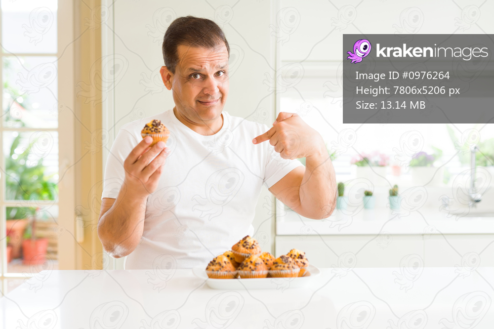 Middle age man eating chocolate chip muffins at home with surprise face pointing finger to himself