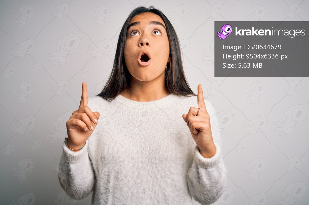 Young beautiful asian woman wearing casual sweater standing over white background amazed and surprised looking up and pointing with fingers and raised arms.