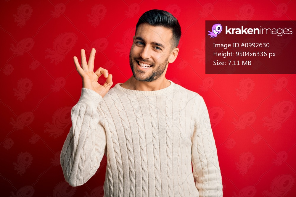 Young handsome man wearing casual white sweater standing over isolated red background smiling positive doing ok sign with hand and fingers. Successful expression.