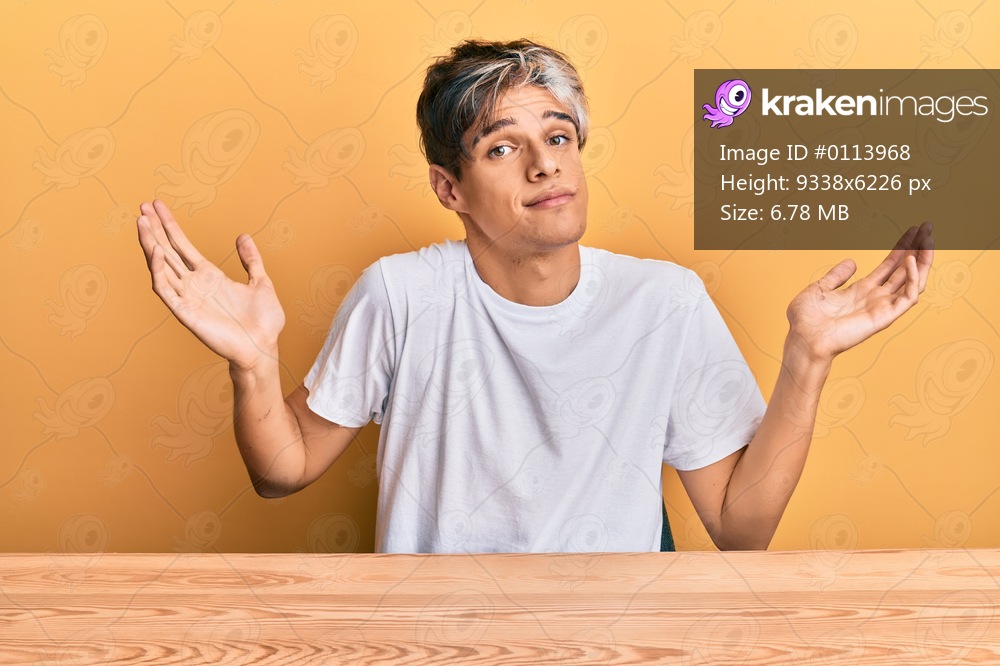Young hispanic man wearing casual clothes sitting on the table clueless and confused with open arms, no idea and doubtful face. 