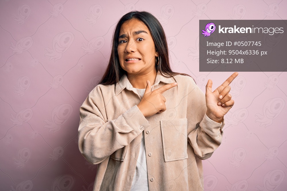 Young beautiful asian woman wearing casual shirt standing over pink background Pointing aside worried and nervous with both hands, concerned and surprised expression